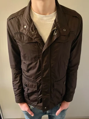 Brun vindjacka från Massimo Dutti - Stilren brun vindjacka från Massimo Dutti med flera fickor framtill och knappdetaljer. Jackan har dragkedja och tryckknappar, samt en klassisk krage. Materialet är lätt och passar perfekt för blåsiga dagar. Snygg och enkel att matcha till olika outfits.