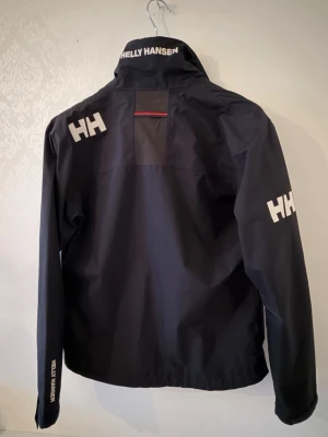 Svart vindjacka Helly Hansen - Snygg svart vindjacka från Helly Hansen med vita HH-loggor på ärm och axel samt röda detaljer på dragkedjorna. Perfekt för blåsiga dagar och sportig stil. Använd få tal gånger. Topp skick 
