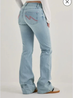 Ljusblå bootcut jeans med brodyr - Säljer ett par ljusblå bootcut jeans med rosa zigzag-broderi på bakfickorna. Jeansen har hög midja och klassisk femficksdesign. Perfekta för en avslappnad och trendig look. De är bootcut