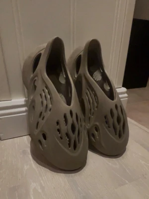 Beige Adidas Yeezy Foam Runner tofflor - Säljer ett par beigea Adidas Yeezy Foam Runner tofflor med futuristisk design och många ventilationshål. Tillverkade i ett lätt och flexibelt skummaterial som ger grym komfort. Perfekta för dig som vill ha något unikt och trendigt på fötterna.