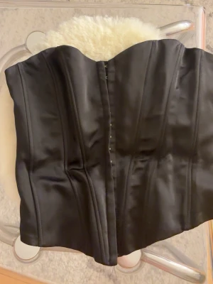 Svart korsett från Zara - Svart axelbandslös korsett från Zara i satinmaterial. Figurnära modell med markerad midja och hakknäppning framtill. Perfekt för en stilren och trendig look.💕