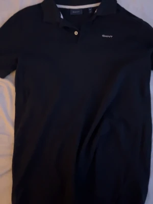 Svart pikétröja från GANT - Klassisk svart pikétröja från GANT med vit logga broderad på bröstet. Tröjan har krage, knappar framtill och korta ärmar. Perfekt till jeans eller shorts för en clean och stilren look.