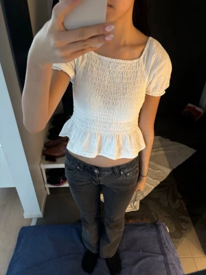 Vit smockad topp med volangkant - Söt vit topp med smockad struktur, korta puffärmar och volangkant nedtill. Toppen har en fyrkantig ringning fram och djupare rygg med knappar. Tillverkad i ett lätt, texturerat tyg som känns luftigt och somrigt. Sitter som XS ungefär💓