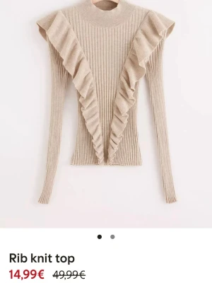 Beige ribbad stickad topp med volanger - Trendig beige ribbstickad topp med långa ärmar och höghalsad design. Volangdetaljer framtill och vid axlarna ger en cool och unik look. Perfekt att styla till jeans eller kjol för en snygg outfit.