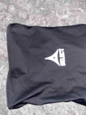 Svart Select sporthalsduk - Svart sporthalsduk från Select med logotyp framtill. Tillverkad i ett tunt och stretchigt syntetmaterial som är perfekt för träning och utomhusaktiviteter. Halsduken har en enkel design och passar bra under jackan eller tröjan när det är kyligt.