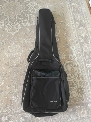 Svart gitarrväska från Gewa - Praktisk svart gitarrväska från Gewa med stor ytterficka och bärhandtag. Väskan är tillverkad i slitstarkt tygmaterial och har vadderad insida för extra skydd. Perfekt för att bära och förvara din gitarr. Enkel och stilren design med dragkedja.
