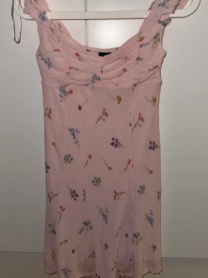 Ljusrosa blommig kortklänning - Söt ljusrosa kortklänning med små blommiga mönster i flera färger. Klänningen har breda axelband och lätt rynkad byst. Tillverkad i ett mjukt, ribbat material som ger en bekväm passform. Perfekt för varma dagar och somriga tillfällen.