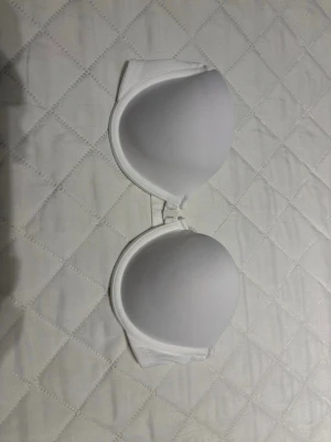 Vit axelbandslös bh med vaddering - Säljer en stilren vit axelbandslös bh med släta, vadderade kupor. Bh:n har en enkel design utan spets eller mönster och är tillverkad i ett glansigt, mjukt material som ger en bekväm känsla. Perfekt under toppar och klänningar med bara axlar.