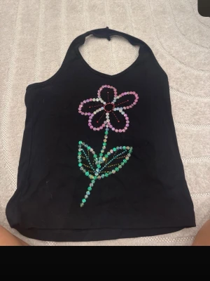 Svart halterneck linnetopp med pärlblomma - Säljer en svart halterneck linnetopp med en stor blomma framtill gjord av färgglada pärlor i rosa, grönt och rött. Toppen är ärmlös och har en enkel, croppad passform som ger en trendig look. Perfekt för dig som gillar unika detaljer och vill sticka ut.