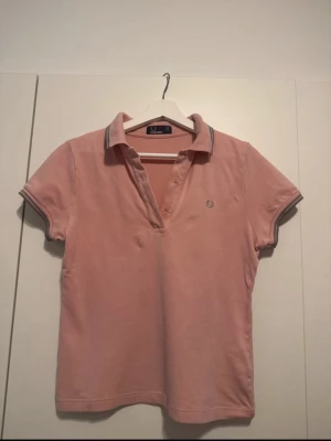 T-shirt fredperry - Säljer en ljusrosa pikétröja från Fred Perry med klassisk krage och knappar framtill. Tröjan har korta ärmar och tunna grå ränder på krage och ärmslut. Tillverkad i mjuk bomull för en bekväm känsla.