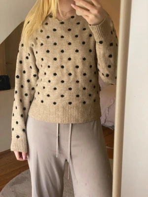 Beige stickad prickig tröja - Mysig beige stickad tröja med svarta prickar och ribbade muddar vid ärmslut och nederkant. Tröjan har en avslappnad passform och rund halsringning. Perfekt för dig som gillar en söt och enkel stil.