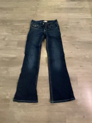 Mörkblå bootcut jeans  - Säljer ett par mörkblå bootcut jeans från Gina Tricot med snygga kontrastsömmar och slitningar framtill. Jeansen har fem fickor, dekorativa knappar på bakfickorna och klassisk låg midja. Perfekta för dig som gillar retrovibbar och vill ha ett par jeans med lite vidare ben. 