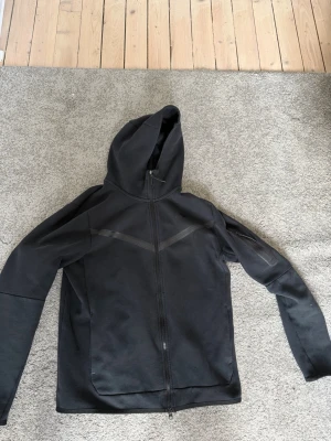 Svart hoodie från Nike med dragkedja - Säljer en svart hoodie från Nike i storlek M. Hoodien har hel dragkedja framtill, två sidofickor och en klassisk huva. Materialet är mjukt och bekvämt, perfekt för chill dagar. Diskreta detaljer över bröstet ger en sportig look.