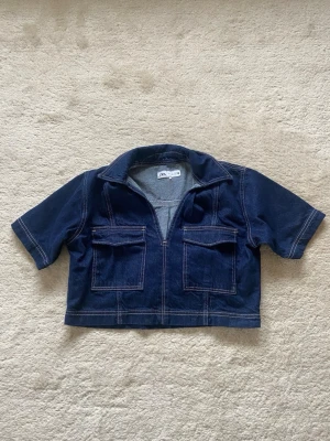 Mörkblå kortärmad jeanstopp från Zara - Snygg mörkblå croppad jeanstopp från Zara med korta ärmar och två stora bröstfickor framtill. Jackan har en öppen krage och markerade sömmar i kontrastfärg. Perfekt för en trendig och avslappnad look.