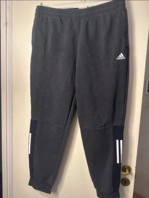 Mörkgråa Adidas träningsbyxor - Mörkgråa träningsbyxor från Adidas med vita ränder på nedre delen av benen och Adidas-logga på vänster lår. Byxorna har resår i midjan och muddar vid bensluten. Perfekta för gymmet eller chill hemma.