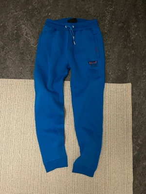 Blåa mjukisbyxor från Puma - Snygga blåa mjukisbyxor från Puma med dragsko i midjan och ribbade muddar vid bensluten. Byxorna har två sidofickor och Puma-logga broderad på vänster ben. Perfekta för chill eller träning.