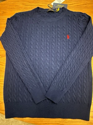 Mörkblå kabelstickad tröja Polo Ralph Lauren - Mörkblå kabelstickad tröja från Polo Ralph Lauren med rund halsringning och röd broderad logga på bröstet. Tröjan har långa ärmar och ribbade muddar vid ärmslut och nederkant. Perfekt för en klassisk och stilren look. Säljer den för den var för liten för mig fick den i julklapp