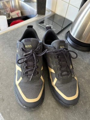Limited Edtion Svarta sneakers från HUGO BOSS - Snygga svarta sneakers från BOSS med beige detaljer och svart sula. Skorna har en sportig design med mesh- och textilmaterial, samt klassisk snörning. Perfekta för dig som vill ha en stilren och modern look. Skorna har även en del i reflex material som ger en snyggare look under mörkare klimat.