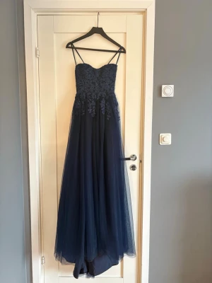 Marinblå långklänning med spetsdetaljer - Elegant marinblå långklänning med tunna axelband och vacker spets upptill. Kjolen är i flera lager tunn tyll som ger ett svepande intryck. Klänningen har snörning i ryggen för justerbar passform . Säljer pågrund av att den är lite för liten och inte riktigt passade mig. Jag och även hon som jag köpte den av är 165 och klänningen funkar bäst med 5 cm klack. Inläggen är uttagna.