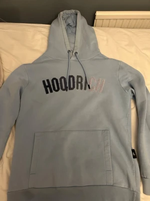 Ljusblå hoodie från Hoodrich - Ljusblå hoodie från Hoodrich med stor magficka och en liten ficka i ärmen. broderad logga i svart och vitt på bröstet.  Perfekt för dig som gillar streetwear och vill ha en chill look. Nypris cirka 900