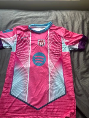 Rosa Barcelona matchtröja Nike - Säljer en Barcelona matchtröja i rosa och ljusblått med vita grafiska linjer och klubbmärke på bröstet. Tröjan har Spotify-logga framtill, lila detaljer på ärmarna och blåprickig krage. Tillverkad i polyester och elastan, perfekt för fotboll eller träning.