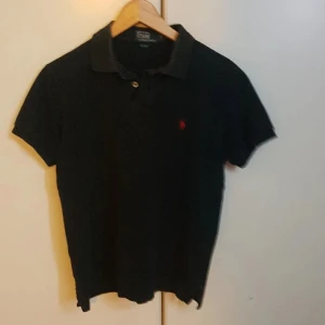 Svart pikétröja från Polo Ralph Lauren - Klassisk svart pikétröja från Polo Ralph Lauren med korta ärmar och krage. Tröjan har en liten röd broderad logga på bröstet och knäppning vid halsen. Perfekt till jeans eller shorts för en clean och stilren look.