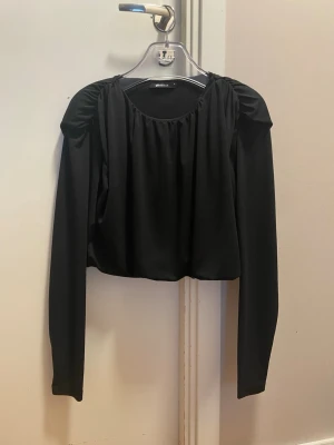 Svart croppad långärmad topp - Snygg svart croppad topp från Gina med långa ärmar och rynkade axlar. Perfekt för att lyfta din outfit med en enkel men trendig look. Mjuk och följsam känsla i materialet.