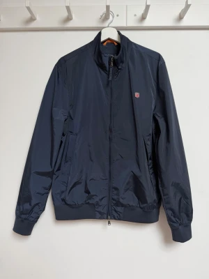 Mörkblå vårjacka från Morris - Stilren mörkblå vindjacka från Napapijri med hög krage, dragkedja framtill och två sidofickor. Jackan har ett litet broderat logomärke på bröstet och elastiska muddar vid ärmslut och midja. Tillverkad i lätt, glansigt material som står emot vind. Nypris 2995 kr. Nyskick!