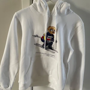 Vit hoodie Polo Ralph Lauren björn - Vit hoodie från Polo Ralph Lauren med ikonisk Polo Bear-tryck där björnen åker skidor. Hoodien har huva med dragsko och en klassisk känguruficka framtill. Mjuk bomullsmix och ribbade muddar vid ärmslut och nederkant. Perfekt för en chill och sportig look.