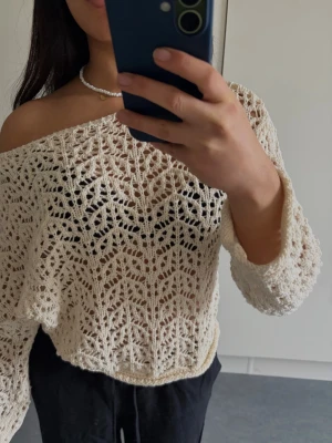 Off shoulder virkad tröja cremevit - Trendig cremevit virkad tröja med off shoulder-design och vida ärmar. Tröjan har ett detaljerat spetsmönster och är gjord i ett mjukt, luftigt garn. Perfekt för lager på lager och ger en avslappnad men snygg look.