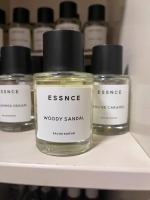 Essnce Woody Sandal Eau de Parfum - Woody Sandal från Essnce är en Eau de Parfum med en transparent, ljusgul vätska i en rund glasflaska med svart cylinderformad kork. Minimalistisk vit etikett med svart text. Perfekt för dig som gillar träiga och sandelträdoftande parfymer.