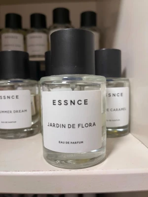ESSNCE Jardin de Flora parfym - Minimalistisk parfymflaska från ESSNCE med doften Jardin de Flora. Flaskan är genomskinlig med en vit etikett och svart text, samt svart cylinderformad kork. Eau de Parfum-varianten ger en stilren och modern känsla, perfekt för dig som gillar enkla och snygga detaljer.