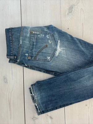 Blå slitna jeans från Dondup - Säljer ett par blå jeans från Dondup med slitningar och distressed detaljer på bakfickorna och benen. Jeansen har raka ben och klassisk femficksdesign. Perfekta för en avslappnad och trendig look.