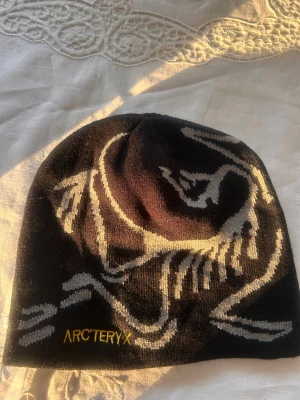 Svart mössa från Arc'teryx - Säljer en svart mössa från Arc'teryx med grått och vitt mönster samt broderad logga i gult framtill. Mössan är tunn och smidig, perfekt för kyliga dagar. Materialet känns mjukt och stretchigt, vilket gör den bekväm att bära.