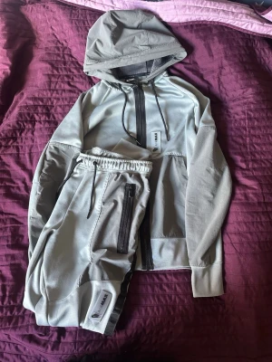 Grått Nike Air Max loungewear-set - Snyggt grått loungewear-set från Nike Air Max med huva och dragkedja på tröjan samt matchande byxor med resårmidja och dragkedjeförsedda fickor. Setet har svarta detaljer och är tillverkat i mjukt, stretchigt material för maximal komfort.