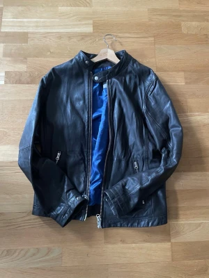 Brun skinnjacka med blått foder - Snygg brun skinnjacka med blått innerfoder och ståkrage. Jackan har dragkedja framtill, två fickor med dragkedja och tryckknappar vid ärmslut och krage. Perfekt för dig som vill ha en stilren och klassisk look. Köpt på Volt ord pris 3000kr