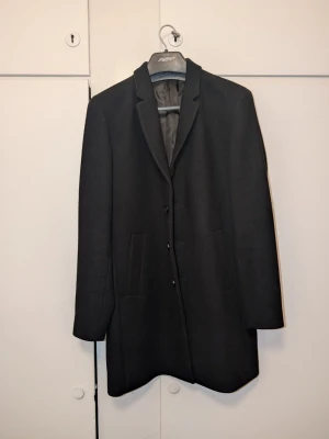 Svart rock från Jack & Jones - Stilren kort rock från Jack & Jones i storlek Large. Tre knappar framtill och diskreta sidofickor. Rocken har en slät ytterväv och är helfodrad med ett grått innerfoder med mönster. Den är endast använd två gånger och är i mycket fint skick. Det bakre skyddsstygnet (slitsen) är fortfarande intakt. Mannen på bilden är 187 cm lång.