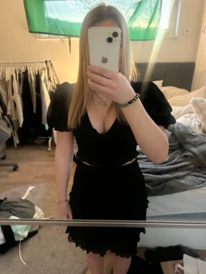 Svart kortklänning med puffärmar - Svart kortklänning med v-ringning och puffiga kortärmar. Klänningen har volangdetaljer och små cut-outs vid midjan. Materialet ser ut att vara polyester och klänningen har en feminin och trendig look som passar perfekt för dig som gillar att sticka ut.