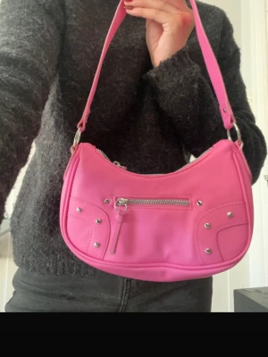 Rosa axelväska med silverdetaljer - Cool rosa handväska i syntetmaterial med silverfärgade nitar och dragkedja framtill. Väskan har en böjd form och justerbar axelrem. Perfekt för dig som gillar färg och vill sticka ut. Ingen synlig logga eller märke.