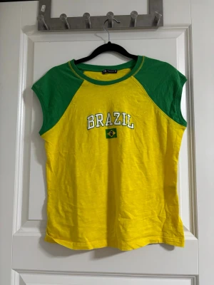 Gul och grön Brazil t-shirt - Gul t-shirt med gröna ärmar och krage. Trycket på bröstet har texten 'BRAZIL' och en liten brasiliansk flagga. T-shirten är ärmlös och har en sportig vibe, perfekt för dig som vill sticka ut.