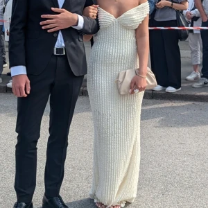 Vit långklänning med öppen rygg - Supersnygg vit långklänning med offshoulder och öppen rygg. Klänningen har en figurnära passform, v-ringad urringning och fina volangdetaljer längs axlarna. Baksidan pryds av en stor rosett och materialet har en strukturerad, nästan pärl-liknande yta. Perfekt för bal eller student! Andvänd 1 gång!  Storlek xxs men passar xxs-S