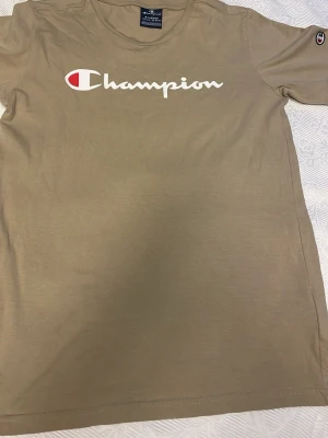 Beige t-shirt från Champion - Snygg beige t-shirt från Champion med stort vitt och rött logotryck över bröstet. Klassisk rund halsringning och korta ärmar. Tillverkad i mjukt bomullsmaterial som känns skönt mot huden. Perfekt för en avslappnad och sportig stil.