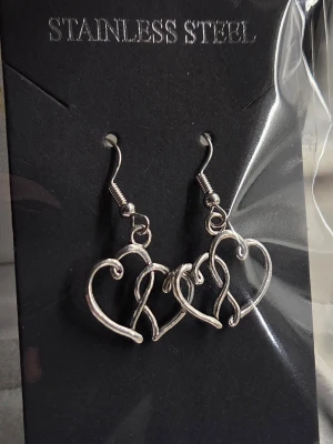 Silvriga hjärtörhängen i stål - Snygga örhängen i rostfritt stål med en öppen hjärtformad design. De har en modern, snirklig look och hängs i örat med krok. Perfekta för dig som gillar stilrena accessoarer med en twist.