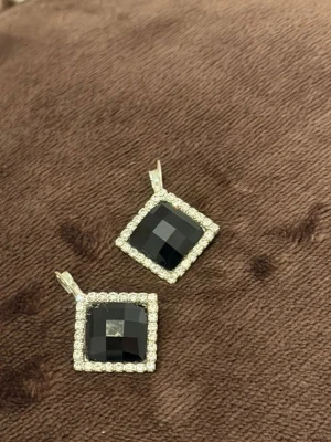 Svartvita fyrkantiga örhängen - Säljer ett par stilrena örhängen med stora, svartslipade stenar i fyrkantig form. Runt stenarna sitter en ram av små, gnistrande vita stenar som ger extra bling. Örhängena har silverfärgade detaljer och en modern look.