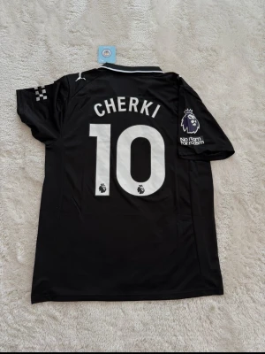 Svart Manchester City tröja Cherki 10 - Snygg svart Manchester City matchtröja från Puma med Cherki och nummer 10 på ryggen. Tröjan har Premier League-märke på ärmen, Etihad Airways-logga på bröstet och klassisk krage med vit detalj. Perfekt för fotbollsfans som vill sticka ut på planen eller läktaren.
