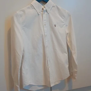 Vit skjorta från Ralph Lauren - Klassisk vit skjorta från Ralph Lauren med button-down krage och broderad logga på bröstet. Skjortan har lång ärm, knappar framtill och är tillverkad i bomull. Perfekt till en clean och stilren look.