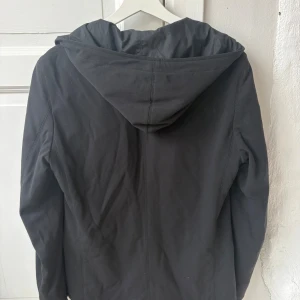 Svart jacka/kavaj Hugo Boss - Snygg svart jacka från Hugo med huva och dragkedja framtill. Jackan har två fickor och en stilren design som passar perfekt till vardagslooken. Materialet känns mjukt och lätt, och modellen har en normal passform, sitter som storlek 46 trots att det är 48