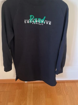 Svart sweatshirtklänning Collective - Cool svart sweatshirtklänning från Collective med tryck framtill i vitt och grönt. Långärmad modell med avslappnad passform och mjukt material, perfekt för en chill och trendig look.