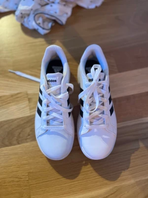 Vita Adidas sneakers med svarta ränder - Snygga vita sneakers från Adidas med de klassiska tre svarta ränderna på sidan. Skorna har vit sula, vita skosnören och en låg profil. Tillverkade i syntetmaterial och textil för en clean och sportig look.