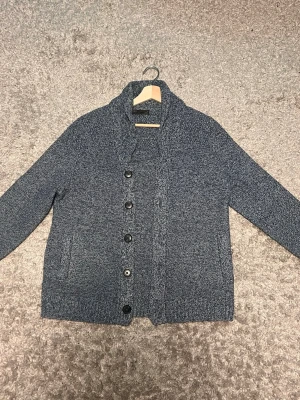 Mörkblå grovstickad kofta - Säljer en mörkblå grovstickad kofta med sjalkrage och stora svarta knappar framtill. Koftan har två fickor på sidorna och är tillverkad i ett mjukt, tjockt material. Enkel men stilren design som funkar till det mesta.